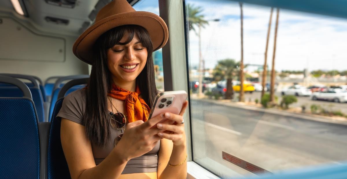 Woman in a hat smiling at her phone on a bus with palm trees outside the window