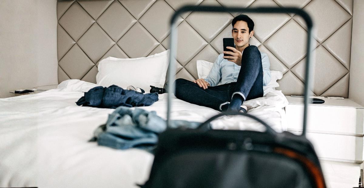 Man sitting on a hotel bed looking at his phone, with a suitcase in the foreground